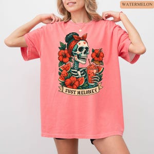May include: A coral-colored t-shirt featuring a skeleton design with a red bandana, surrounded by red hibiscus flowers and watermelon slices. The skeleton is holding a drink with a straw. The text "JUST MELONEY" is on a banner.