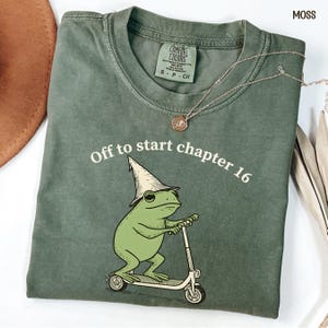 May include: Moss green t-shirt with the text "Off to start chapter 16" and a cartoon frog wearing a wizard hat and riding a scooter. A gold necklace with a pendant is draped across the shirt.