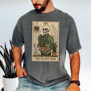 May include: Gray t-shirt featuring a tarot card design with a skeleton holding a potted plant. The card reads "XIV" at the top and "THE PLANT DAD" at the bottom. The design includes other plants and a neutral color palette.