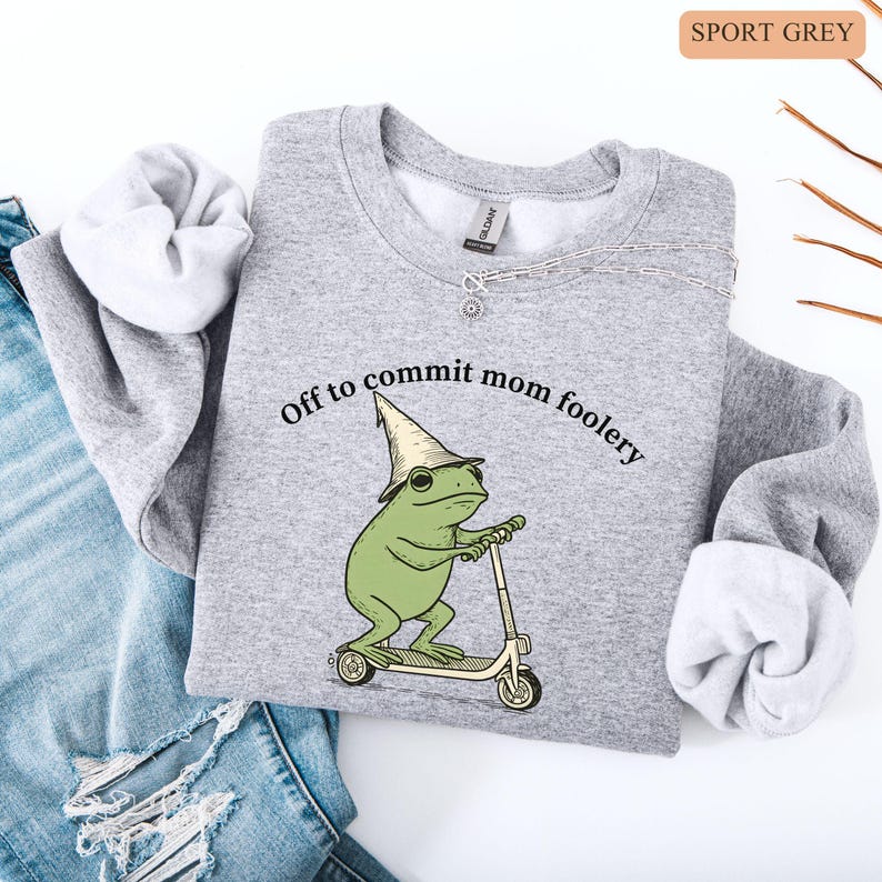 May include: Sport grey sweatshirt with the phrase "Off to commit mom foolery" above a green frog riding a scooter and wearing a wizard hat. The sweatshirt has a crew neck and long sleeves.