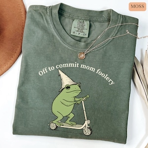 May include: Moss green Comfort Colors t-shirt with the phrase "Off to commit mom foolery" and a cartoon frog riding a scooter. The frog wears a pointed hat. A gold necklace is draped on the shirt.