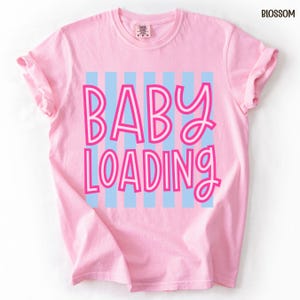 May include: A light pink t-shirt with the words "BABY LOADING" in a pink and blue design. The text is centered on the shirt, with blue vertical stripes behind the text. The shirt has rolled-up sleeves.
