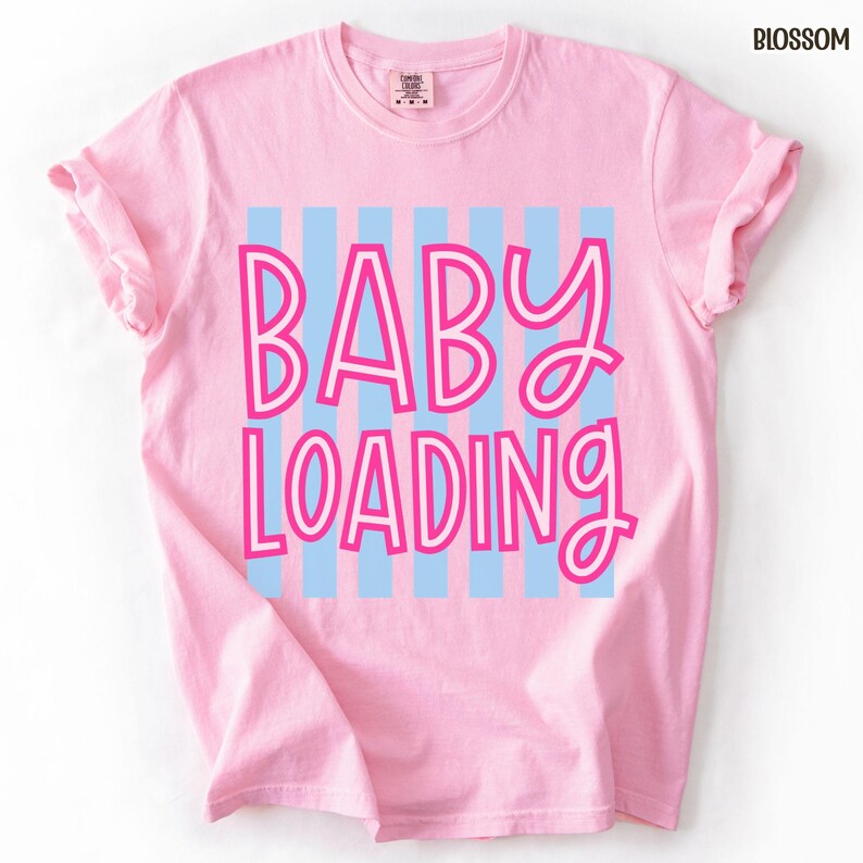 May include: A light pink t-shirt with the words "BABY LOADING" in a pink and blue design. The text is centered on the shirt, with blue vertical stripes behind the text. The shirt has rolled-up sleeves.