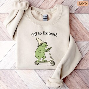 May include: Sand-colored sweatshirt with the text "Off to fix teeth." The graphic features a green frog wearing a wizard hat and riding a scooter. The sweatshirt is made of a soft material.