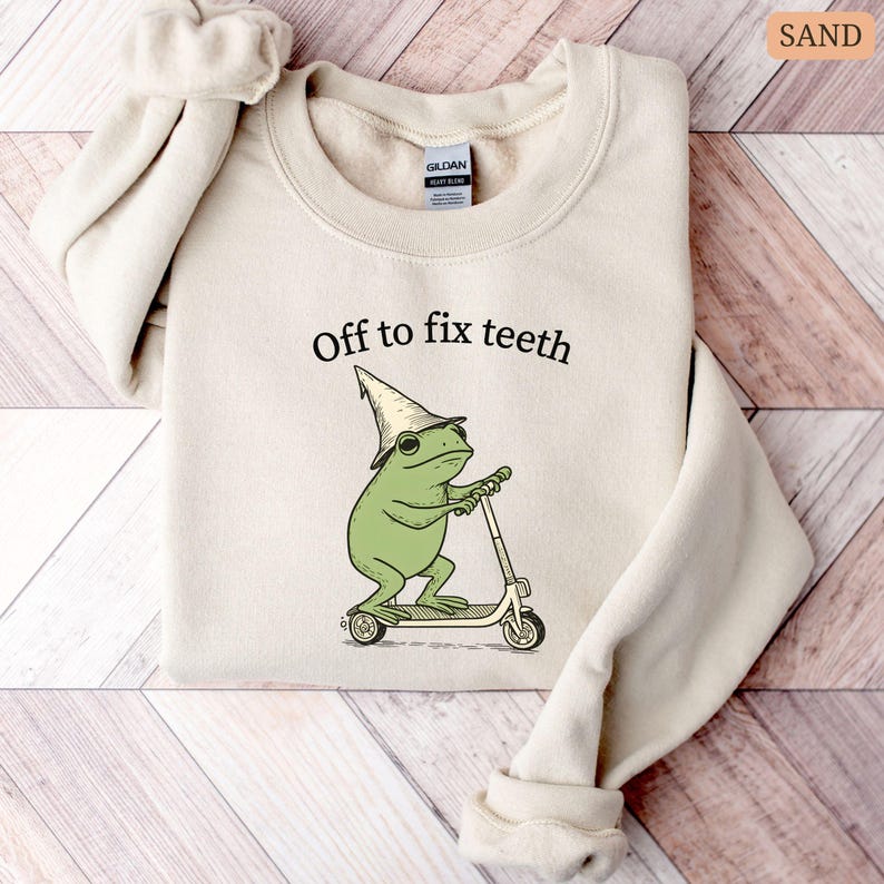 May include: Sand-colored sweatshirt with the text "Off to fix teeth." The graphic features a green frog wearing a wizard hat and riding a scooter. The sweatshirt is made of a soft material.