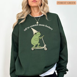 May include: Forest green sweatshirt with the phrase "Off to commit mom foolery" above a cartoon frog riding a scooter. The frog wears a wizard hat. The sweatshirt is a solid color.