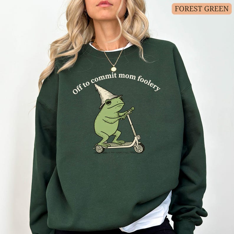 May include: Forest green sweatshirt with the phrase "Off to commit mom foolery" above a cartoon frog riding a scooter. The frog wears a wizard hat. The sweatshirt is a solid color.