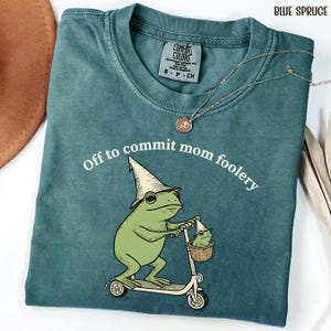 May include: Blue spruce t-shirt with a frog graphic riding a scooter and text that reads "Off to commit mom foolery." The frog wears a wizard hat and carries a smaller frog in a basket. A gold necklace is draped across the shirt.