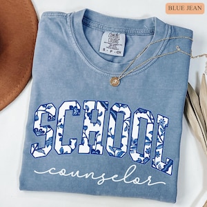 May include: A blue jean Comfort Colors t-shirt with the word "SCHOOL" in large white letters with a blue floral pattern. The word "counselor" is written in cursive below. A gold necklace with a pendant is on the shirt.