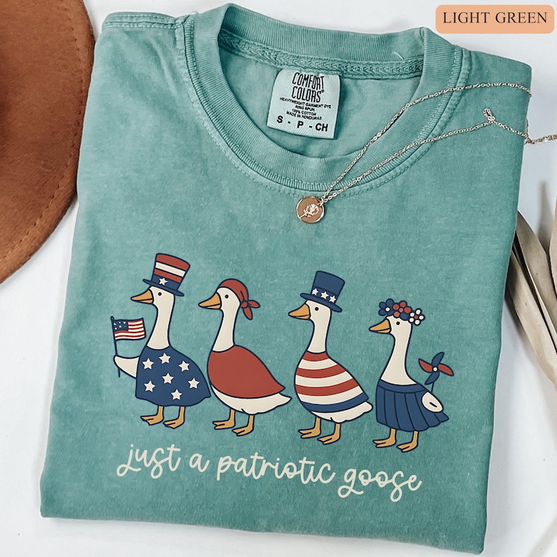 May include: Light green Comfort Colors t-shirt featuring four patriotic geese in festive attire. One goose holds an American flag. The text "just a patriotic goose" is printed at the bottom. A silver necklace is draped across the shirt.