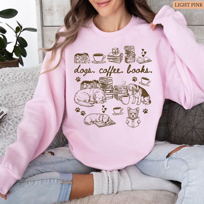 May include: Light pink sweatshirt with a graphic design featuring dogs, coffee cups, books, and paw prints. The text "dogs. coffee. books." is printed in a brown color. The sweatshirt is a crew neck style.