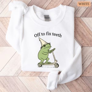 May include: White sweatshirt with the text "Off to fix teeth" above a cartoon frog riding a scooter. The frog is green and wearing a hat. The sweatshirt is folded on a wooden surface.