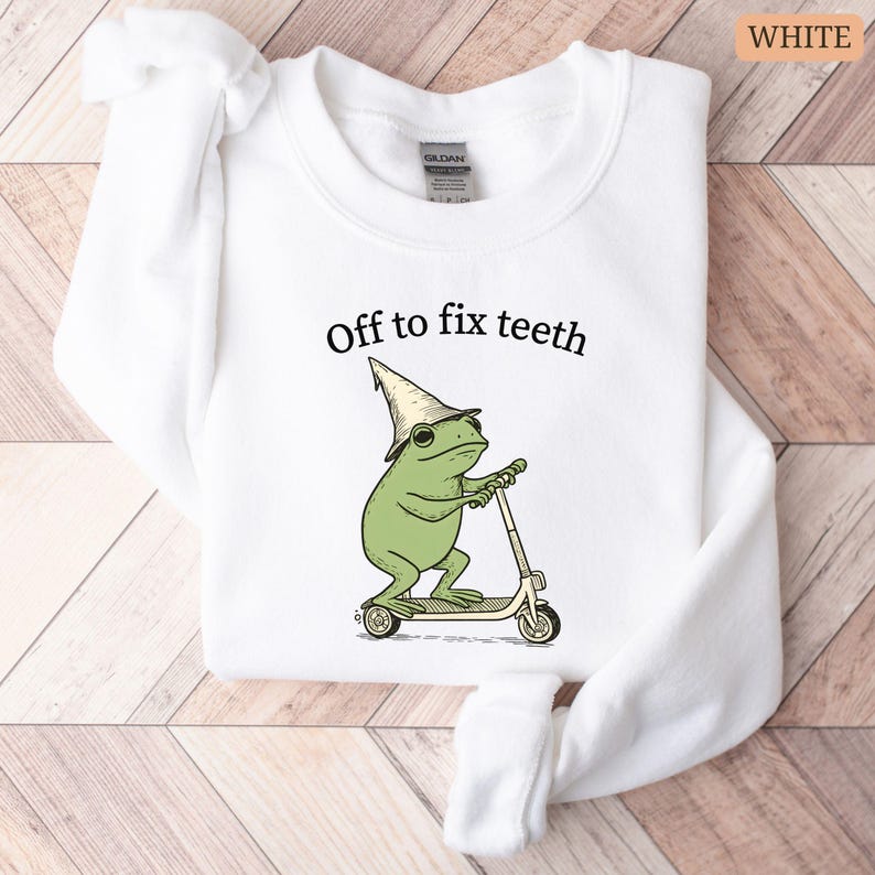 May include: White sweatshirt with the text "Off to fix teeth" above a cartoon frog riding a scooter. The frog is green and wearing a hat. The sweatshirt is folded on a wooden surface.