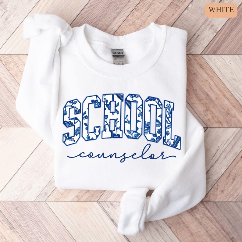 May include: White sweatshirt with the word "SCHOOL" in large, blue floral print letters, and the word "counselor" in a smaller, cursive font. The sweatshirt is folded on a light wood-grain surface.