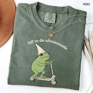 May include: Moss green t-shirt with the text "Off to do ultrasounds" above a cartoon frog riding a scooter and wearing a wizard hat. A silver necklace with a pendant is draped across the shirt.