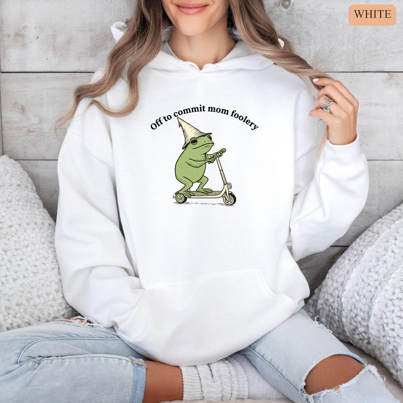 May include: White hoodie with a graphic of a green frog wearing a hat and riding a scooter. The text "Off to commit mom foolery" is printed above the frog. The hoodie has a drawstring hood and a front pocket.