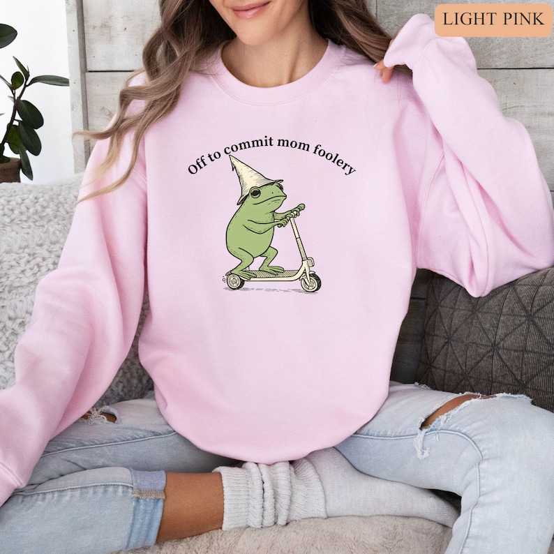 May include: Light pink sweatshirt with the text "Off to commit mom foolery." The graphic features a green frog wearing a wizard hat and sunglasses, riding a scooter. The sweatshirt is a solid light pink color.