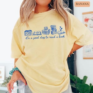May include: A light yellow t-shirt with a blue graphic of books, a coffee cup, and hearts. The text "it's a good day to read a book" is printed below the graphic. The shirt is a relaxed fit.