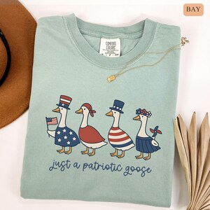 May include: A light green t-shirt featuring a patriotic goose design. Four geese are dressed in red, white, and blue outfits, with one holding a small American flag. The text "just a patriotic goose" is printed below the geese.