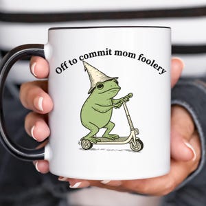 May include: White ceramic mug with a black handle and rim. The mug features a green frog wearing a wizard hat and riding a scooter. Text on the mug reads "Off to commit mom foolery."