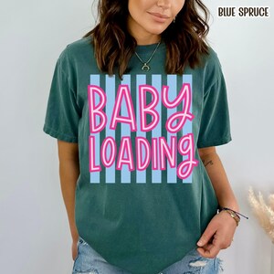 May include: A teal-colored t-shirt with the words "BABY LOADING" in pink lettering outlined in white, set against a light blue striped background. The text "BLUE SPRUCE" is in the upper right corner.