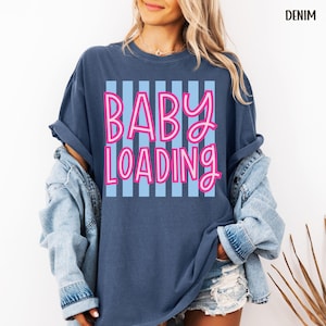 May include: A denim blue t-shirt with the words "BABY LOADING" in pink script over a light blue striped background. The shirt has a relaxed fit and rolled-up sleeves.