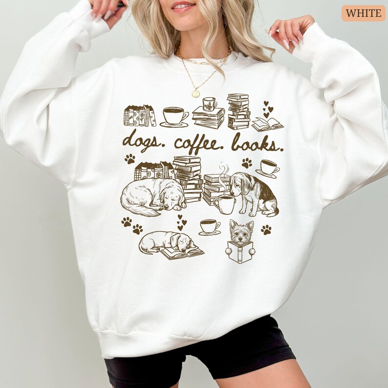 May include: White sweatshirt with a brown graphic featuring dogs, coffee cups, and books. The design includes paw prints, hearts, and the words "dogs. coffee. books." in a whimsical font. The sweatshirt is a casual, comfortable style.