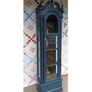 May include: A tall, dark blue, wooden display cabinet with glass doors and shelves. The cabinet has a decorative top with finials and a gold knob. The shelves are lined with a patterned paper.