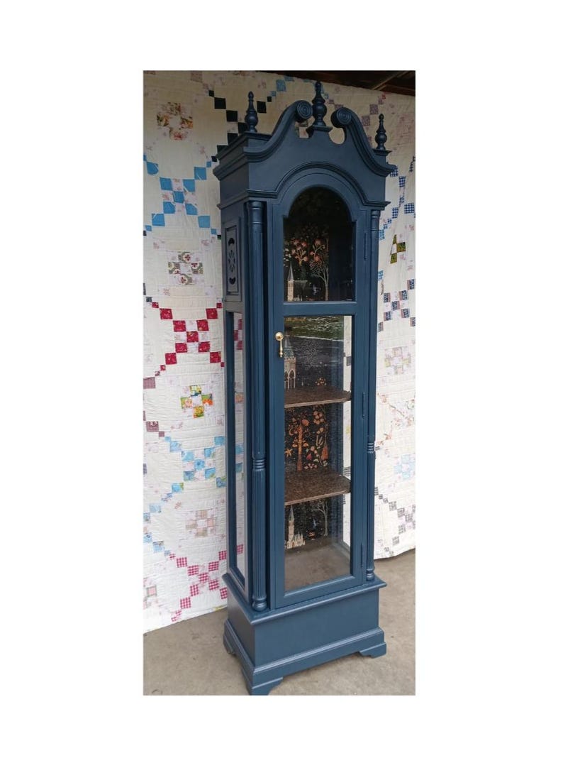 May include: A tall, dark blue, wooden display cabinet with glass doors and shelves. The cabinet has a decorative top with finials and a gold knob. The shelves are lined with a patterned paper.