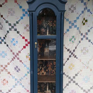 May include: A tall, dark blue wooden cabinet with glass doors and shelves. The cabinet has a decorative top with a curved design and gold accents. The shelves are lined with a black and white patterned paper featuring a whimsical forest scene with trees, houses, and stars.