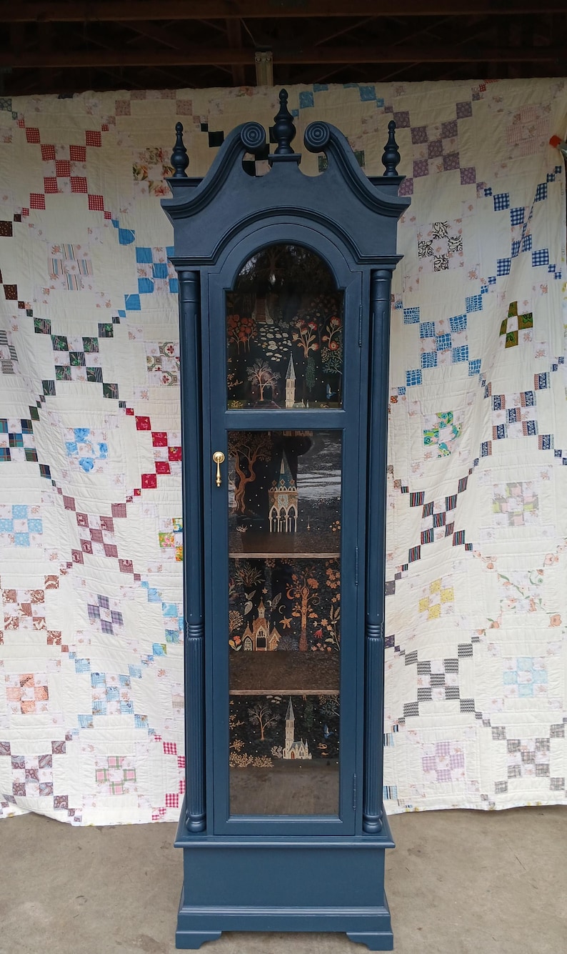 May include: A tall, dark blue wooden cabinet with glass doors and shelves. The cabinet has a decorative top with a curved design and gold accents. The shelves are lined with a black and white patterned paper featuring a whimsical forest scene with trees, houses, and stars.