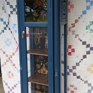 May include: A tall, dark blue wooden display cabinet with glass doors and shelves. The cabinet has a decorative top with ornate details and a gold-colored handle. The shelves are lined with a black and gold patterned paper.