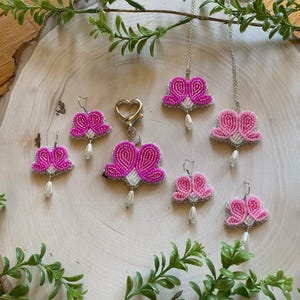 Op de afbeelding: Een collectie handgemaakte kralen sieraden, waaronder oorbellen, een ketting en een sleutelhanger. De stukken hebben een bloemmotief in roze en witte tinten, met een bungelende parel accent. De sieraden worden gepresenteerd op een houten oppervlak met groen.