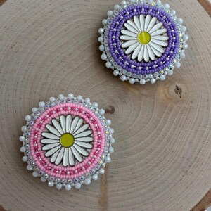 May include: Two circular beaded pins featuring a central white daisy with a yellow center. One pin is bordered by concentric rings of purple beads and clear crystals, while the other is bordered by pink beads and clear crystals, both with white edges.