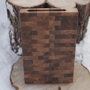 May include: A rectangular wooden chopping board with a dark brown, mosaic-like pattern. The board has a handle cut-out at the top and is set against a snowy backdrop, resting on a tree stump.