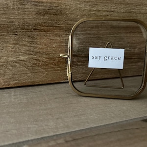 May include: A square, gold-toned metal picture frame with a small white card inside that reads "say grace." The frame is propped up by a small, gold-toned metal triangle. The background is a wooden surface.