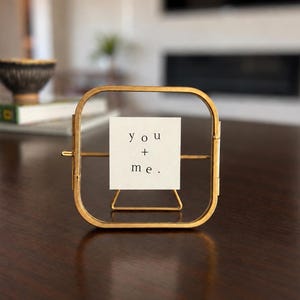 May include: A square gold-toned metal frame with rounded corners, featuring a central glass pane that holds a white square card. The card displays the black printed text "you + me." The frame is supported by a thin metal stand attached to the back.
