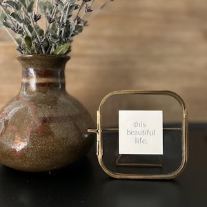 May include: A gold-toned metal picture frame with a glass front and a white card inside. The card has the text "this beautiful life." The frame is sitting on a black surface next to a brown vase with purple flowers.
