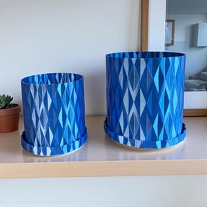 May include: Two blue geometric patterned plant pots with matching saucers. The pots have a diamond pattern in shades of blue and white. The pots are cylindrical and sit on a light-colored surface.