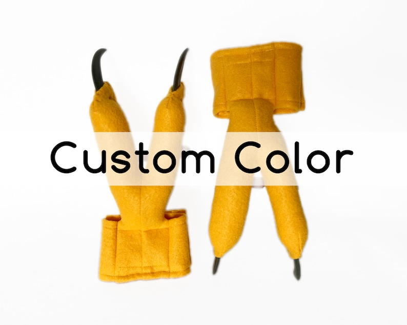 Pode incluir: Um par de garras de dinossauro de feltro amarelo com garras pretas. As garras s&atilde;o presas a uma bra&ccedil;adeira de feltro amarelo. O texto "Custom Color" &eacute; impresso em um fundo branco.