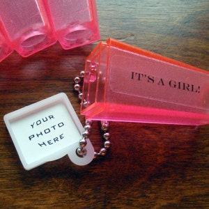 Può includere: Portachiavi in plastica rosa con una cornice bianca che dice "YOUR PHOTO HERE" e un ciondolo in plastica rosa che dice "IT'S A GIRL!"