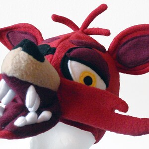 Puede incluir: Disfraz de cabeza de zorro de felpa roja con un ojo blanco y amarillo, dientes afilados y una nariz burdeos.