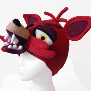 Puede incluir: Disfraz de cabeza de Foxy el Zorro Pirata de felpa roja con dientes blancos y un ojo amarillo. El disfraz se lleva en una cabeza de maniqu&iacute; blanca.