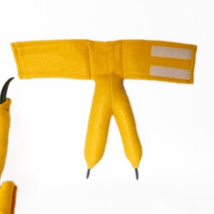 Pode incluir: Um traje de dinossauro de feltro amarelo com uma tira de velcro para prender. O traje tem duas garras longas e uma cauda.