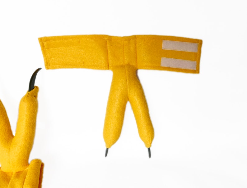 Pode incluir: Um traje de dinossauro de feltro amarelo com uma tira de velcro para prender. O traje tem duas garras longas e uma cauda.