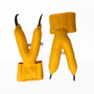 Pode incluir: Um par de garras de dinossauro de feltro amarelo com pontas pretas. As garras s&atilde;o presas a uma faixa de feltro amarelo que pode ser usada no pulso.