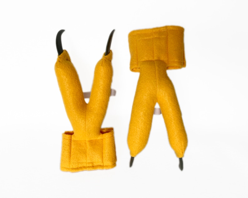 Pode incluir: Um par de garras de dinossauro de feltro amarelo com pontas pretas. As garras s&atilde;o presas a uma faixa de feltro amarelo que pode ser usada no pulso.