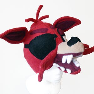 Puede incluir: Sombrero de cabeza de zorro de felpa roja con parche en el ojo. El sombrero tiene una boca grande abierta con dientes blancos.