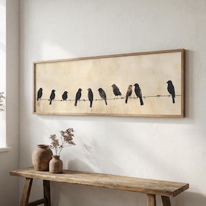 May include: A framed artwork showing black bird silhouettes on a wire against a beige backdrop. The artwork is mounted on a white wall above a wooden bench, with decorative vases. The frame is made of wood.
