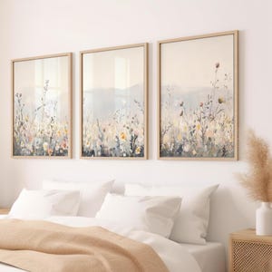 May include: Three framed art prints depicting a watercolour meadow scene. The artwork uses a soft, muted colour palette with beige, yellow, and grey tones. The frames are light wood, creating a calming and natural aesthetic, perfect for a bedroom.